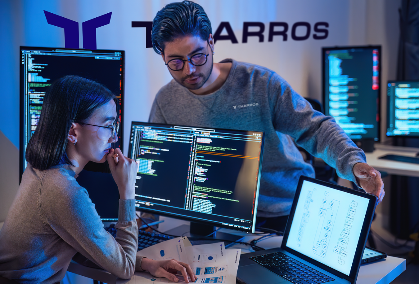 A man wearing glasses and a grey sweatshirt and a woman wearing a grey top collaborate on projects on their computers